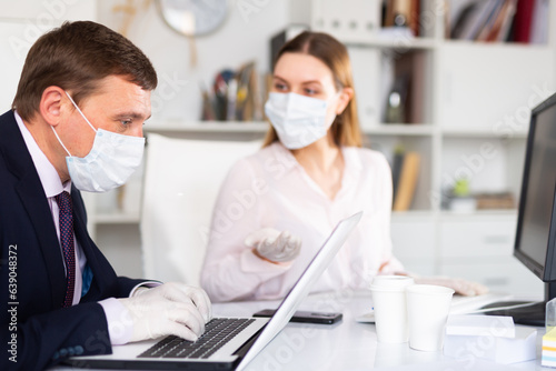 Portrait of busy entrepreneur in medical face mask and latex gloves working with female coworker in office. Concept of precautions and social distancing in coronavirus pandemic..