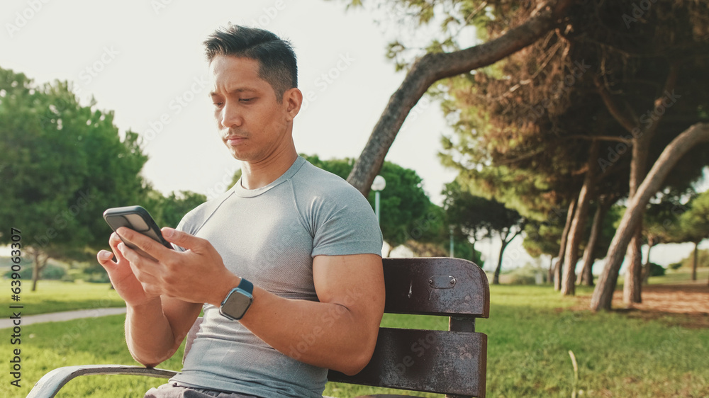 Obraz premium Guy in sportswear, an athlete sits on a bench in a city park, scrolls information, watches videos, writes in social networks on a mobile phone on a sunny day