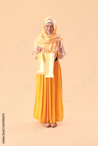Young beautiful woman in hijab opening shopping bags on pale orange background