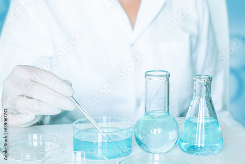 Laboratory scene: Lab assistant conducts procedures. Wearing gloves and a white robe. Employing glassware. Clear blue liquid.