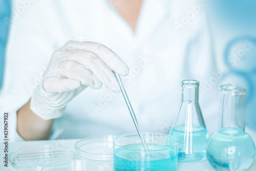 Scientific scene: Lab assistant performs tasks. Gloves and white robe worn. Glass equipment used. Clear blue liquid in play.