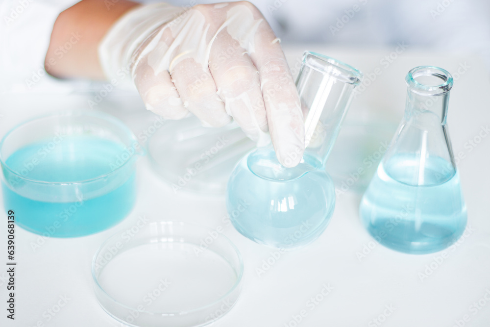 Scientific setting: Lab assistant manipulates liquid in flask. Wearing gloves, white robe, employing glassware. Clear blue liquid involved. Scientific setting: Lab assistant manipulates liquid in flask. Wearing gloves, white robe, employing glassware. Clear blue liquid involved.