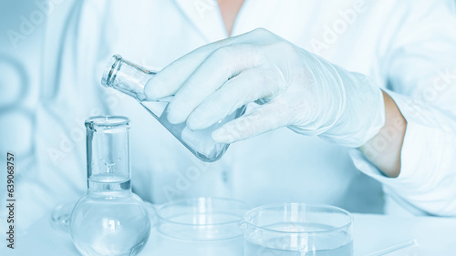Scientific scene: Assistant engages in manipulations in the laboratory setting. Lab coat attire. Glass tools used. Situated in a blue laboratory. Researching with transparent liquid.