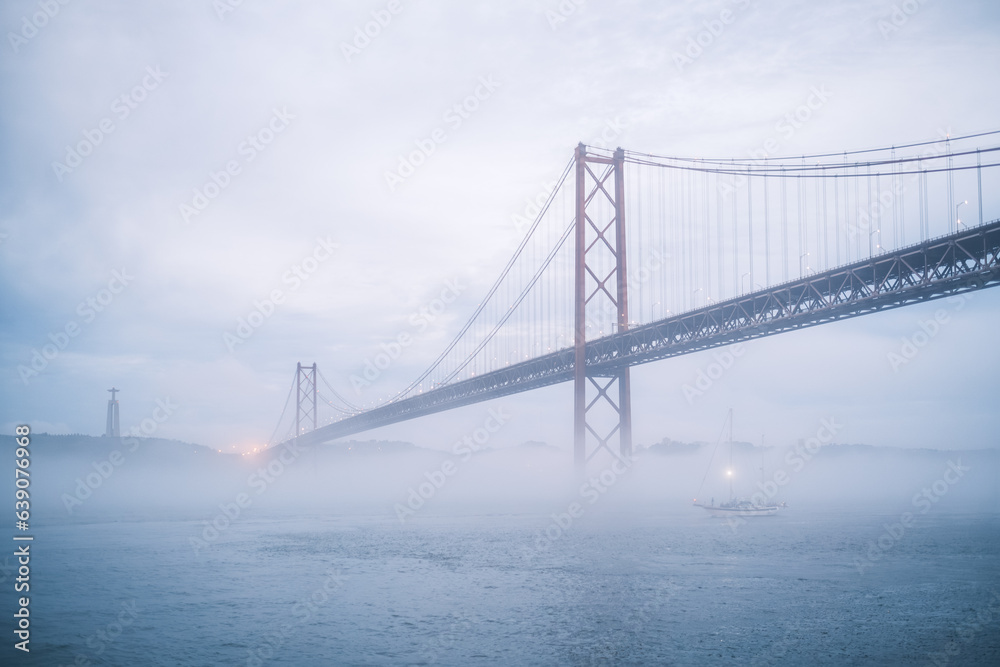 View of 25 de Abril Bridge famous tourist landmark of Lisbon connecting Lisboa and Almada in heavy fog mist wtih yacht boats passing under. Lisbon, Portugal