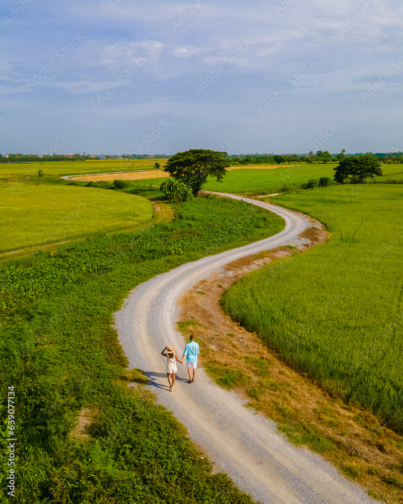 a couple of men and women on vacation in Thailand walking on a curved winding countryside road in the middle of green rice paddy fields in Central Thailand Suphanburi region, drone aerial view of