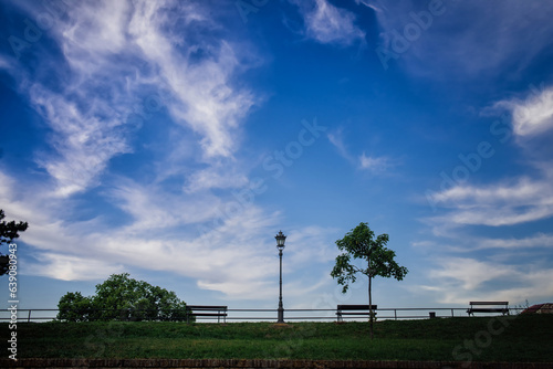 Side view of an alley with green bushes, a lamppost bench against the background of a huge blue sky with unusual beautiful clouds. Minimalistic style, space for text
