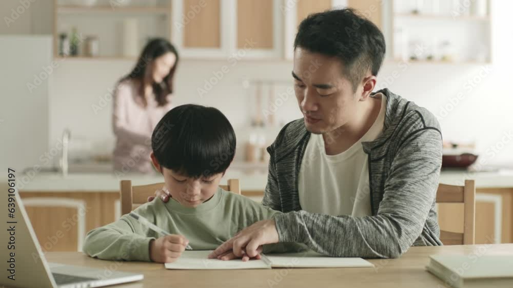 young asian father helping son with homework while mother preparing meal in kitchen young asian father helping son with homework while mother preparing meal in kitchen
