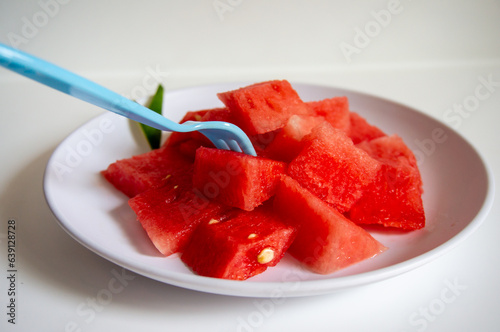 Fresh watermelon ready to eat using a blue fork on a white plate with a white background