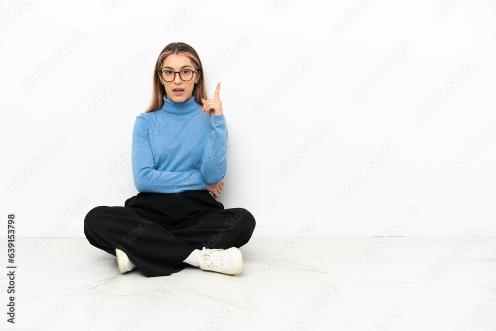 Young Caucasian woman sitting on the floor thinking an idea pointing the finger up