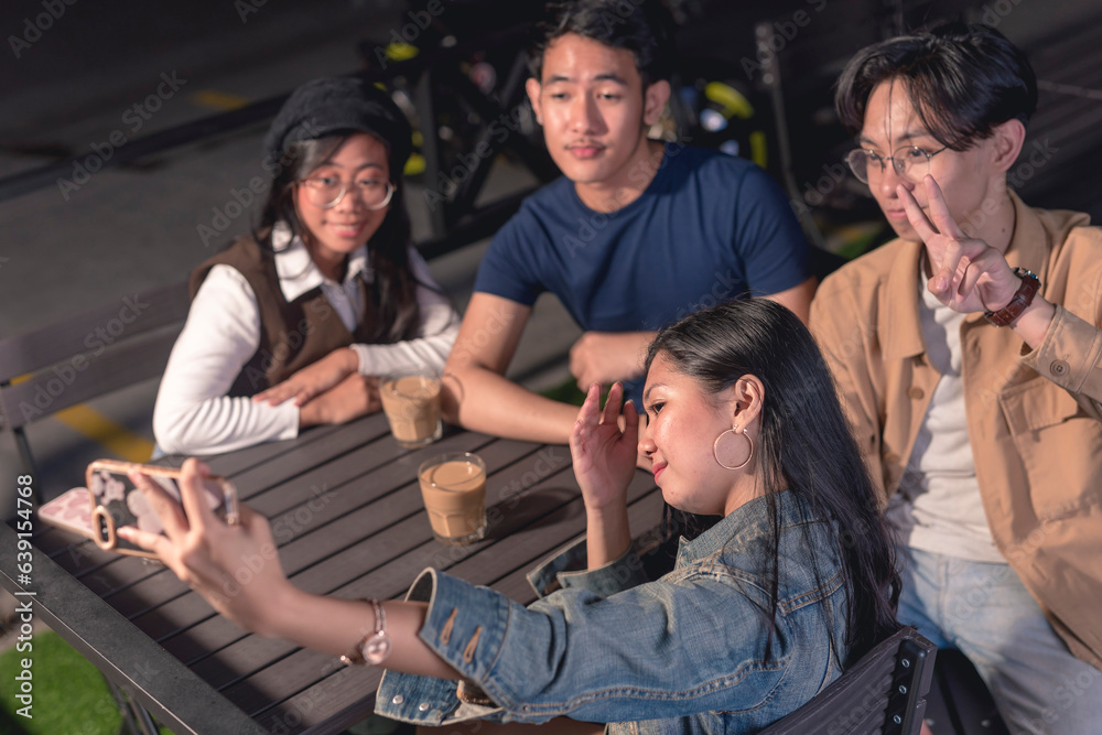 The lady in a denim jacket is holding up a phone to take a picture of herself and with her friends, two of them are ladies and two guys. Motorcycle, parking lot and rail in the background