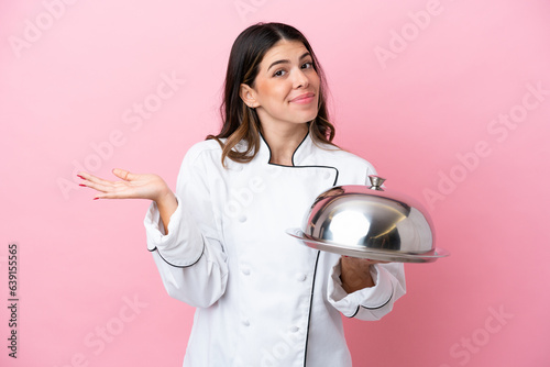 Young Italian chef woman holding tray with lid isolated on pink background having doubts while raising hands photo