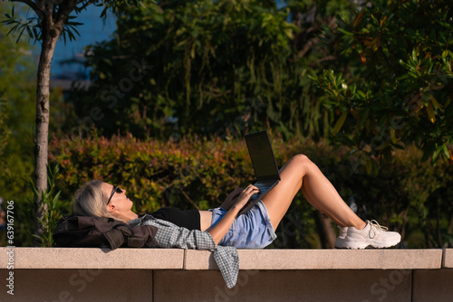 Young slender blonde woman is lying on a park bench with a laptop on a sunny day, side view. Happy girl with a laptop in the park