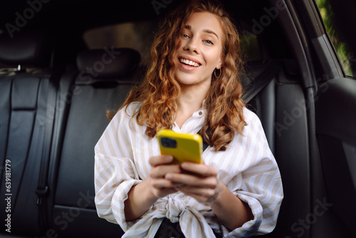 Modern woman in casual clothes sits in the back seat of a car, looks away, uses a mobile phone. Business, technology and travel concept.