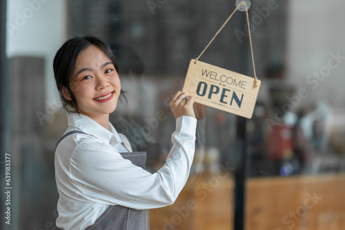 Young freelance businesswoman Asian young female barista in apron holding sign preparing to open for sale and standing in front of the door of the cafe with open sign business owner startup concept