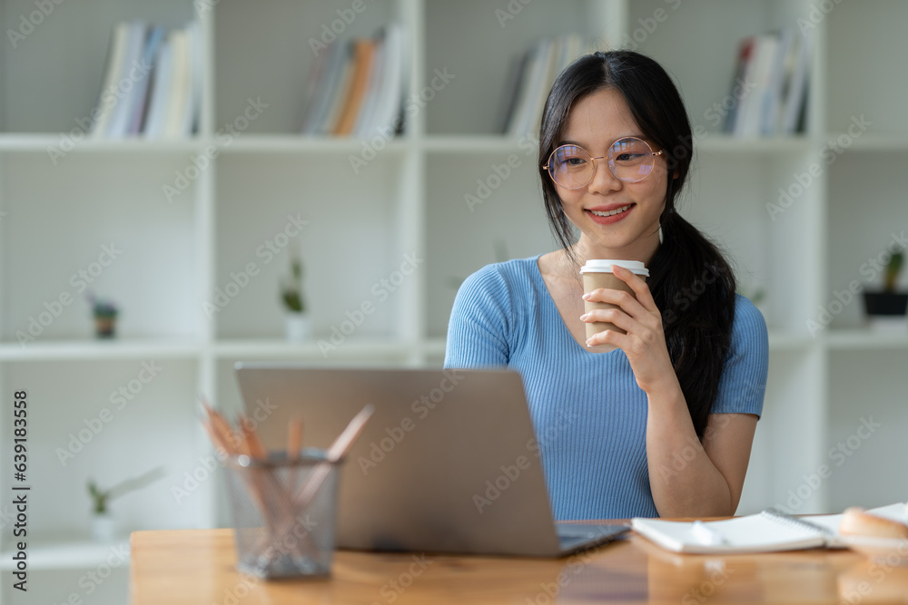 Asian woman sitting happily at work smiling relaxing and chatting with friends on social media search for information on laptop take notes in a notebook while sitting on a desk in his office at home Asian woman sitting happily at work smiling relaxing and chatting with friends on social media search for information on laptop take notes in a notebook while sitting on a desk in his office at home