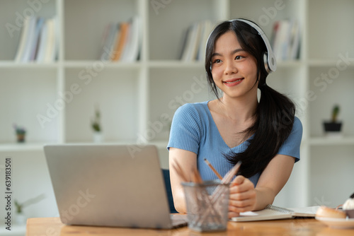 Asian woman wearing headphones sitting and working Listen to relaxing music and chat with friends on social media on laptop while sitting on weekends home office desk.