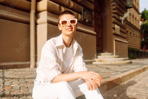 Hipster fashion woman in stylish glasses sits on a curb in the street in Europe, enjoying the sunny weather. Urban street fashion concept.