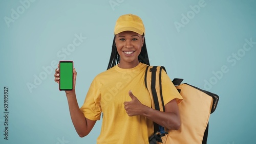 Delivery woman in yellow uniform with portable backpack refrigerator and smartphone shows thumbs up. Advertising area, workspace mockup. photo