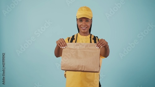 Delivery woman in yellow uniform with portable backpack refrigerator giving paper bag at the camera. Isolated on blue background. photo