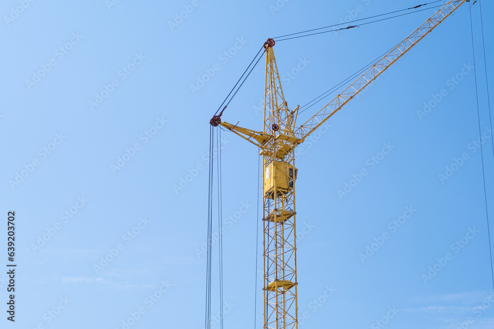 A crane and a building under construction against a blue sky background. Builders work on large construction sites, and there are many cranes working in the field of new construction. A crane and a building under construction against a blue sky background. Builders work on large construction sites, and there are many cranes working in the field of new construction.