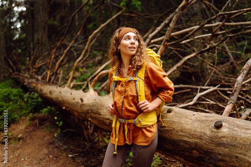  Traveler with a yellow backpack walks along a hiking path among the mountains in the forest. Curly woman tourist enjoys nature  explores new places.