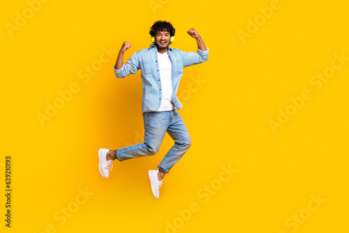 Full length photo of cheerful lucky man wear jeans shirt jumping rising fists enjoying music empty space isolated yellow color background