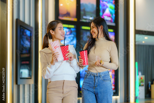 Asian friends walk in front of the cinema holding glasses and popcorn having fun.