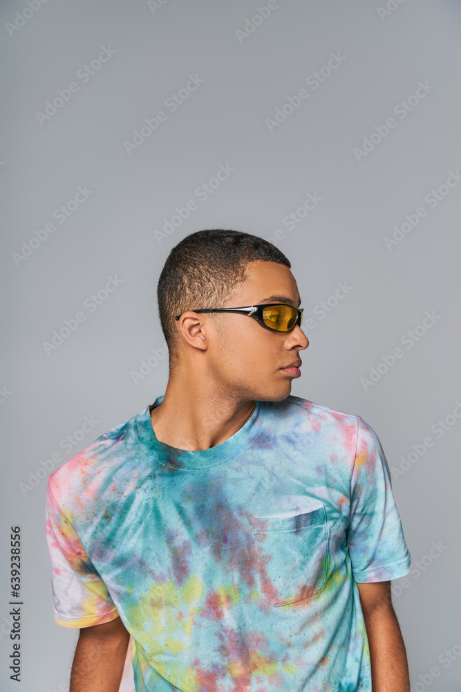 youthful african american man in sunglasses and tie-dye t-shirt looking away on grey youthful african american man in sunglasses and tie-dye t-shirt looking away on grey