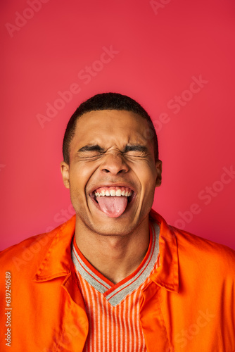funny african american with closed eyes sticking out tongue on red background, orange shirt, stylish