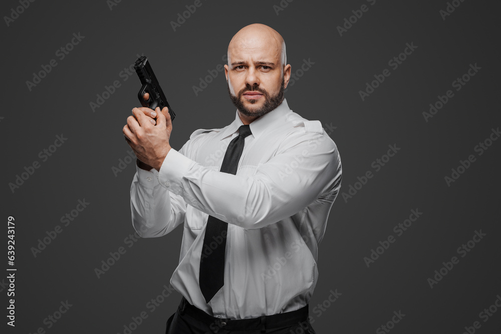 Bald, bearded man in white shirt and tie holds a pistol, epitomizing a detective, bodyguard, or security professional against a gray studio background Bald, bearded man in white shirt and tie holds a pistol, epitomizing a detective, bodyguard, or security professional against a gray studio background
