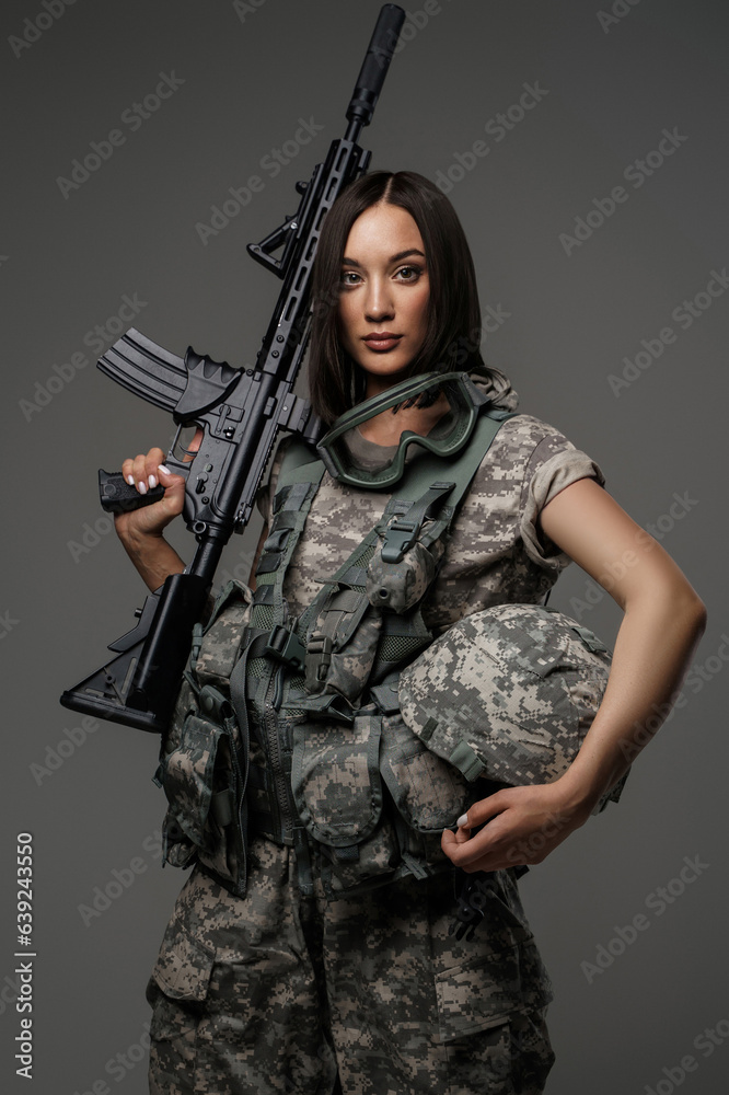 Beautiful female soldier in military uniform stands holding an automatic rifle against a grey background, posing for a stunning studio portrait
