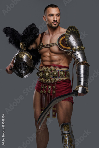 Gladiator with a well-groomed beard and lightweight ornate armor poses with a gladius and feathered helmet against a simple grey backdrop, radiating strength and courage