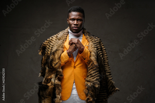 Portrait of a stylish dark-skinned man wearing a yellow jacket and a wild animal skin draped over his shoulders, posing against a dark background