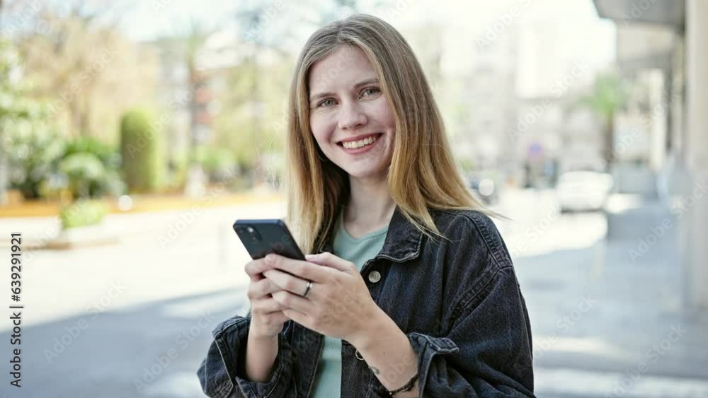 Young blonde woman using smartphone smiling at street