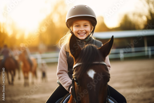 Happy girl kid at equitation lesson looking at camera while riding a horse, wearing horseriding helmet