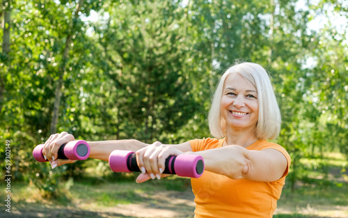 Senior woman going sport exercises with dumbbells on summer park. Active lifestyle concept.