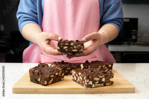 A female pastry chef in a pink apron and blue T-shirt holds a square piece of chocolate cake with both hands