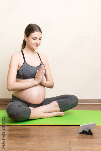 Fototapeta Naklejka Na Ścianę i Meble -  Pregnant female holding smartphone while sitting on exercise yoga mat at home. Fitness or yoga training online at coronavirus time