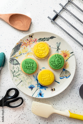 Four cookies on a plate with nature-themed pattern, scissors, kitchen spatula, tongs, and a sieve on a pastry chef's kitchen table