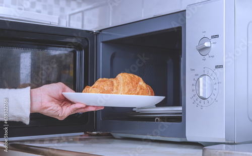 Woman hand putting plate of croissant into microwave oven in kitchen room, close up with copy space