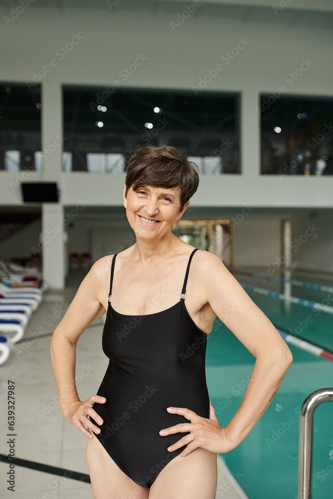 positive middle aged woman standing with hands on hips near swimming pool, swimwear, spa center
