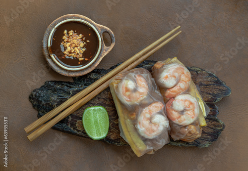Homemade riceberry rice paper spring rolls stuffed with shrimp (prawn), sliced chicken, tofu and egg served with sliced lime and chili sauce sprinkle with ground peanuts on wooden plate. Top view.