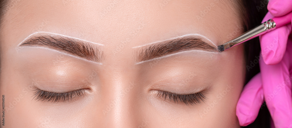 Brunette teenager girl having permanent makeup tattoo on her eyebrows. Make-up artist makes markings with white paste for eyebrow tattooing. Professional makeup and skin care cosmetology.
