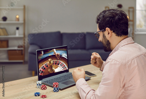 Close-up of young joyful man playing roulette. Lucky guy bets at online casino at home on his laptop. Winning Russian roulette over the Internet on the net. photo