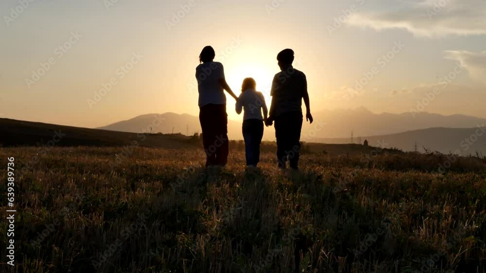 Three children walk through a rural sloping wheat field hand in hand towards the sunset. View from behind teenagers brother and sisters holding hands step into the sunset across the field Three children walk through a rural sloping wheat field hand in hand towards the sunset. View from behind teenagers brother and sisters holding hands step into the sunset across the field