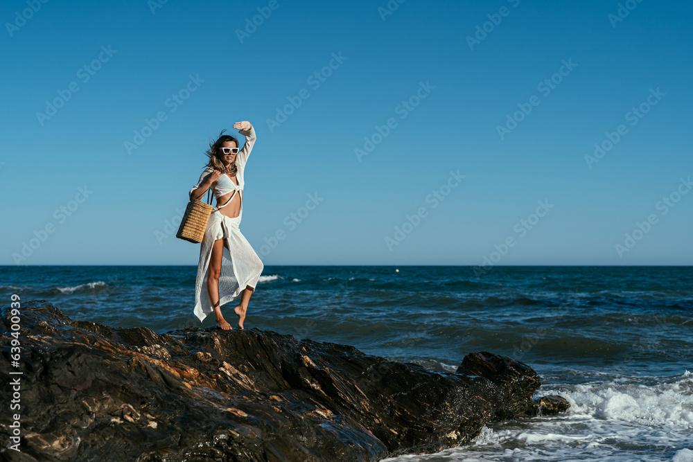 A beautiful girl in white glasses and white clothes with a bag in her hands stands on the stones on the seashore A beautiful girl in white glasses and white clothes with a bag in her hands stands on the stones on the seashore