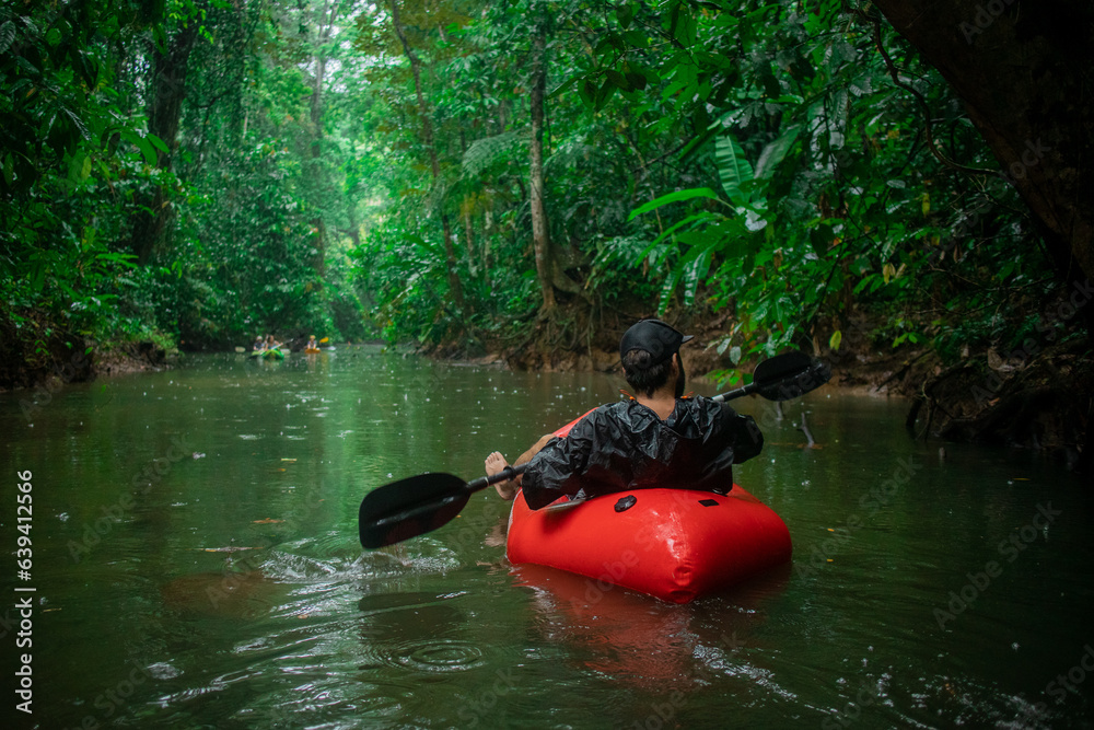 Unknown man in a red kayak floating towards lush vegetation visible around. Palm trees, mangroves, leaves and other plants in rain.