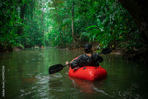 Obraz na płótnie Unknown man in a red kayak floating towards lush vegetation visible around. Palm trees, mangroves, leaves and other plants in rain. w salonie