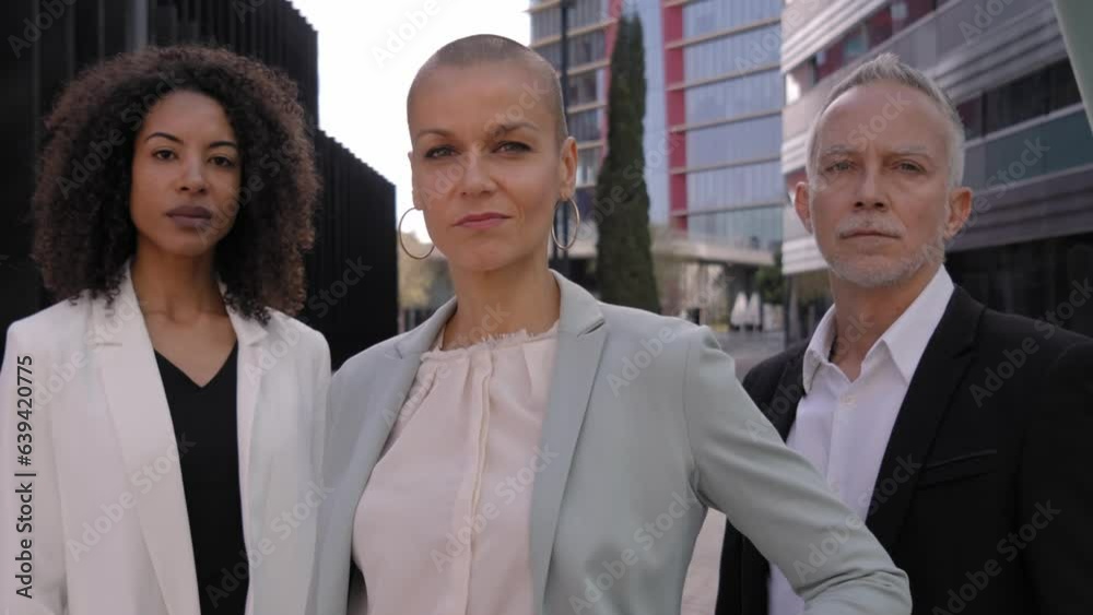 Three multiracial attractive business people pose serious expression outside office center. Diverse work team looking at camera confident standing in city. Professionals formal successful middle-aged. Three multiracial attractive business people pose serious expression outside office center. Diverse work team looking at camera confident standing in city. Professionals formal successful middle-aged.