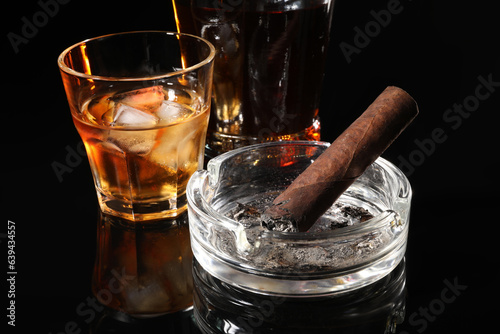 Burnt cigar, ashtray and whiskey on black mirror surface, closeup